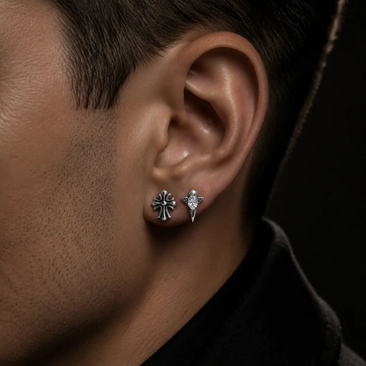 Primer plano de un hombre con dos aretes Gothic plateados distintos en el mismo lóbulo: una cruz y un diseño alado